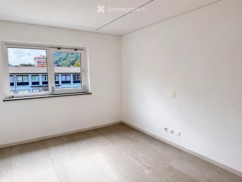 Empty room with light-colored walls, wooden flooring, and a large window offering a view of neighboring buildings and greenery.