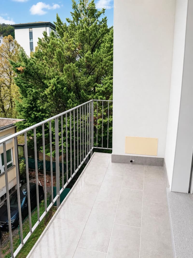 Balcony with grey tiled floor, metal railing, and a view of green trees and neighboring buildings.