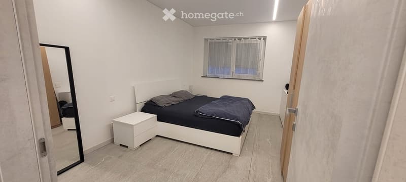 Bedroom: white bed, gray pillows, bed sheet, white night stand, full length mirror, wood floor, window with white curtains