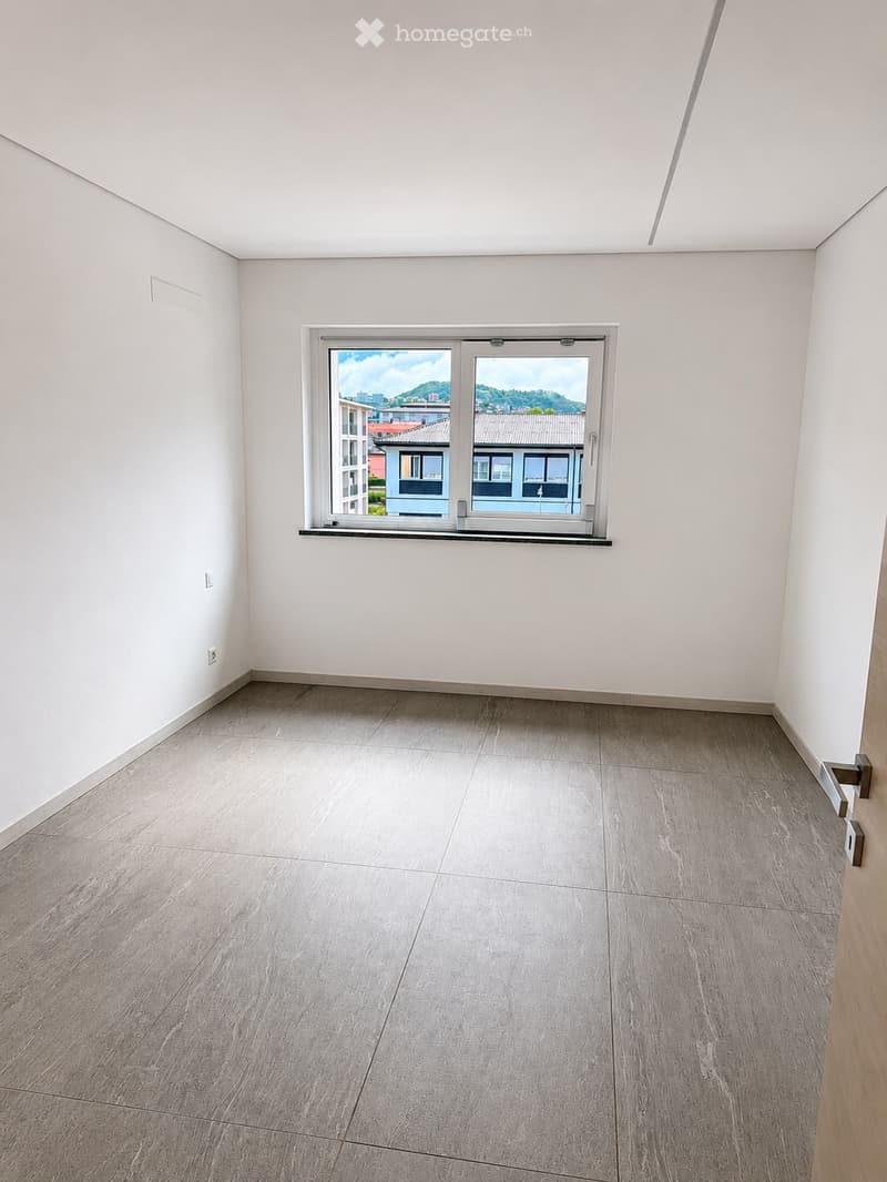 Empty room with light grey tiled floor and white walls, featuring a large window with a view of the city and surrounding landscape.