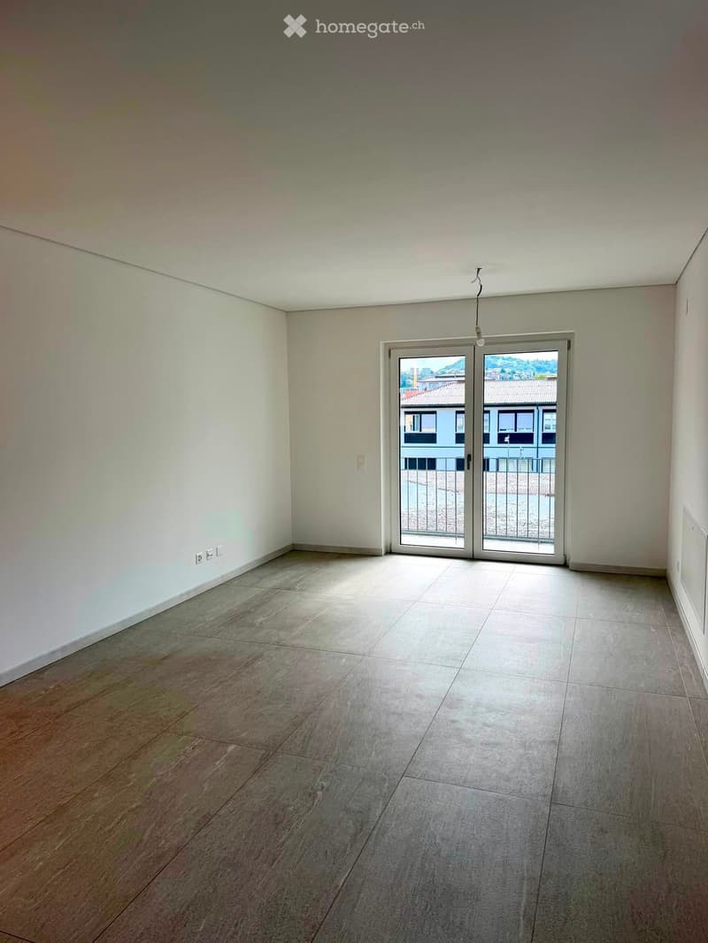 Empty room with light-colored walls, wooden and tiled flooring, and a large sliding glass door leading to a balcony.