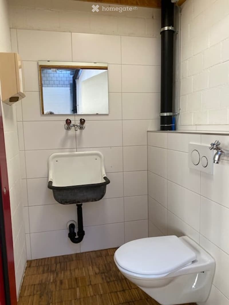 Small bathroom with white tiled walls, wooden floor, toilet, sink, and mirror.