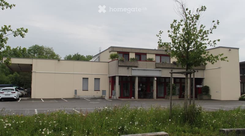 Modern two-story building with beige facade, red-framed windows, and a covered entrance area. The building has a flat roof and is surrounded by a parking lot with several cars. There are small trees and shrubs around the building.