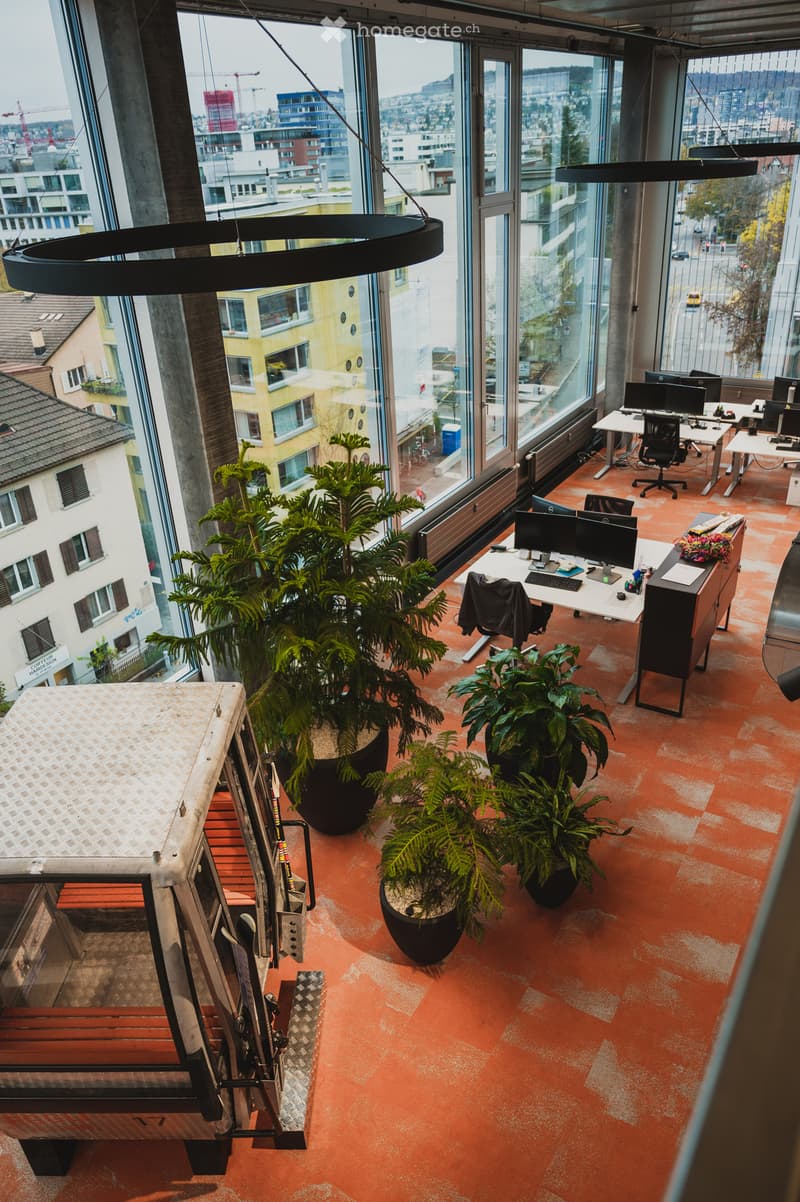 Open floor plan, large windows, desk with monitors, chairs, and potted plants, office vehicle parked