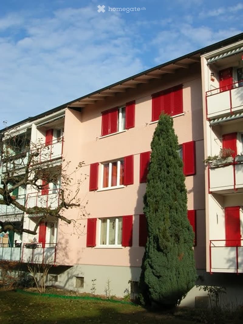 Apartment building with pink exterior and red shutters, multiple balconies, located in a suburban area with greenery and a tree in front.