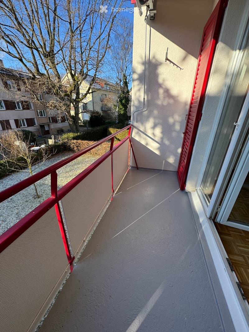 Balcony with red railing, floored with cement, opens to indoor space, glass door with red shutters