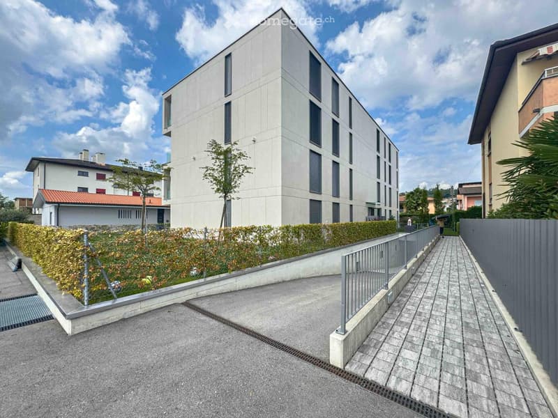 white building with multiple floors, metal fence, paved path, bushes, tree, driveway