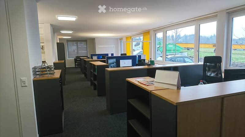 Large open office space with multiple work stations, each having computer monitors, chairs, and storage shelves. Office is equipped with lights on the ceiling, glass windows allowing natural light, and a single power outlet on the wall.