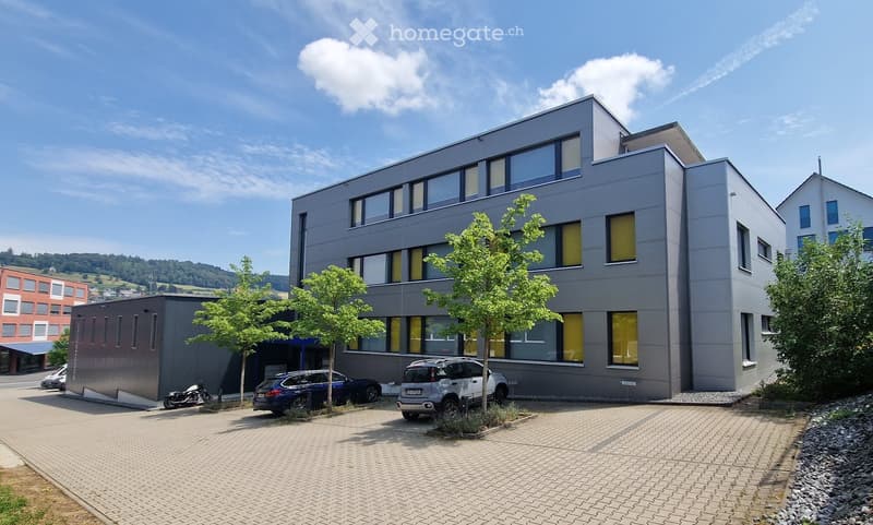 Modern multi-story building with grey facade and large windows, surrounded by trees and parked vehicles.