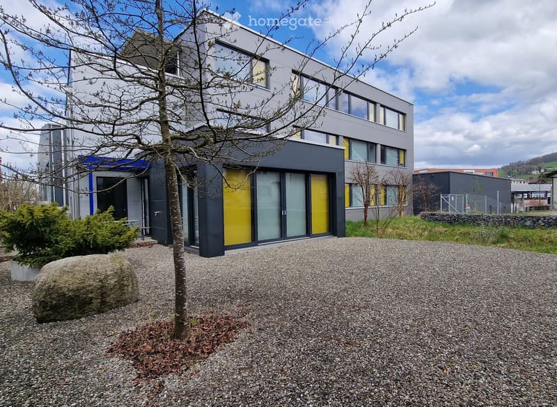 Modern two-story building with grey and yellow facade, large windows, and a gravel driveway.