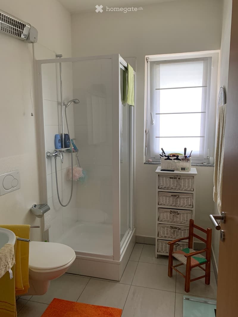 A white tiled bathroom with a glass shower enclosure, a toilet, a white sink, white walls and wooden furniture.