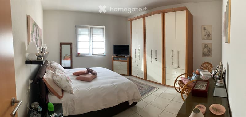 white walls, white tiled floor, large bed with white sheets and pillows, large wardrobe, mirror, TV stand with TV, lamp, desk with framed pictures and a jewelry box, side tables with lamps and figurines