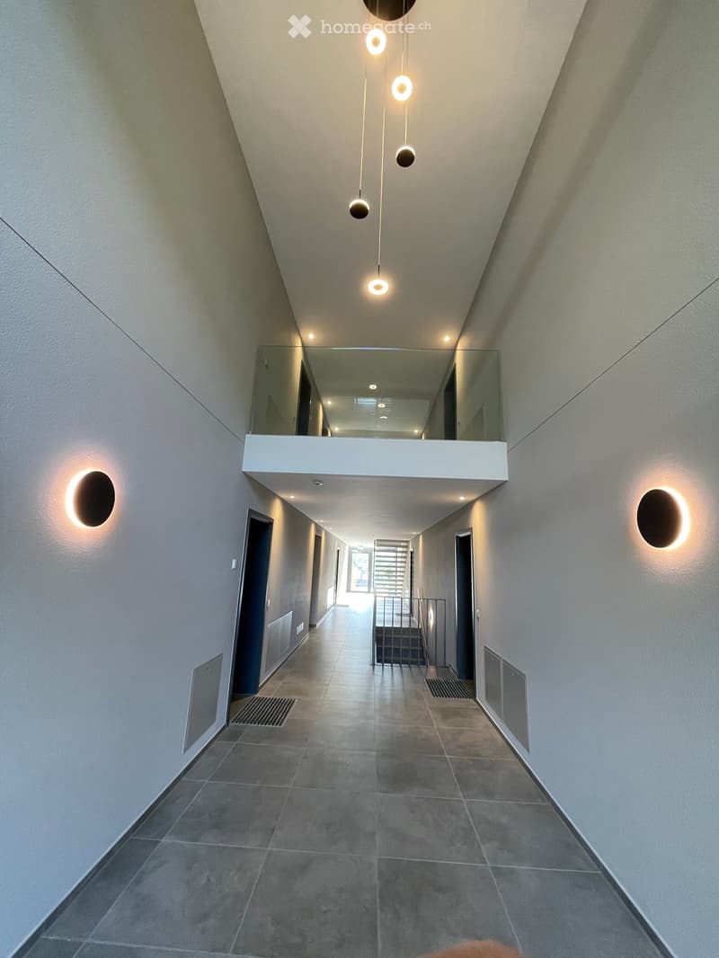 Modern hallway with grey tiled floor, white walls, recessed lighting, and a suspended light fixture with multiple circular bulbs. Features a mirrored balcony overlook and built-in wall-mounted lighting on both sides.