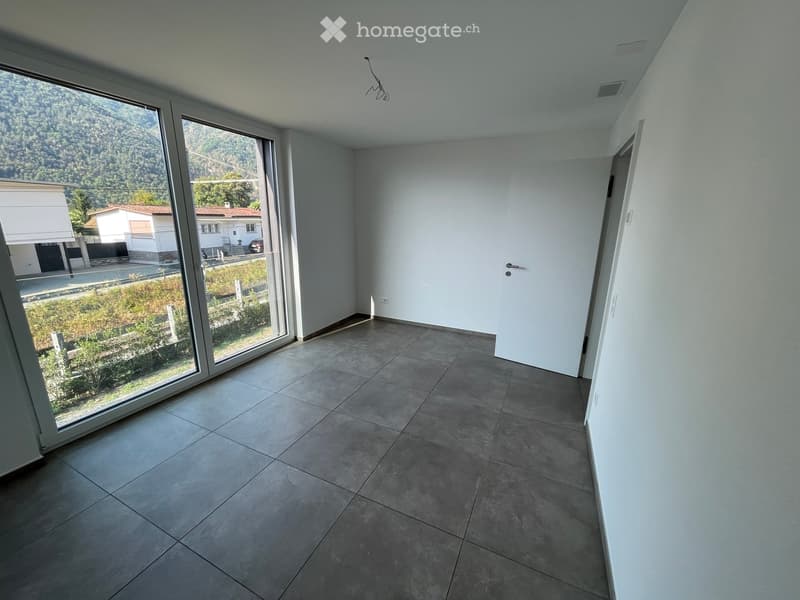 Empty room with grey tiled floor, white walls, large sliding glass door leading to outdoor view, and a closed white door.