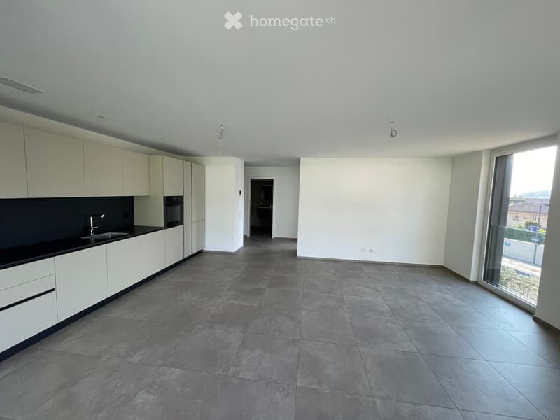 Empty kitchen with white cabinets, black countertop, and large windows, connected to a hallway.