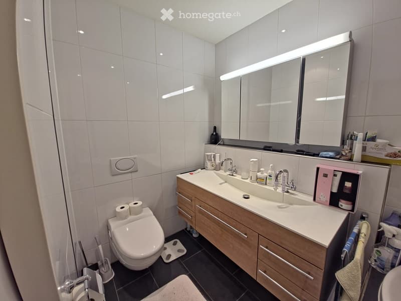 modern bathroom, white tiled walls, white toilet, white sink, wooden vanity, shower area, black tiles on floor, white mat by toilet, mirror above vanity