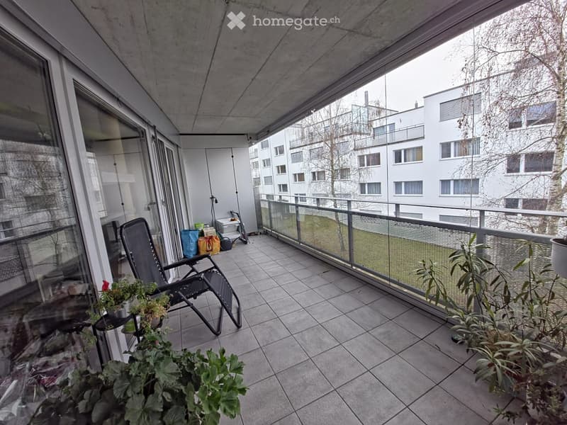 balcony with glass railing, tile floor, 2 black chairs, 3 potted plants, green grass in front