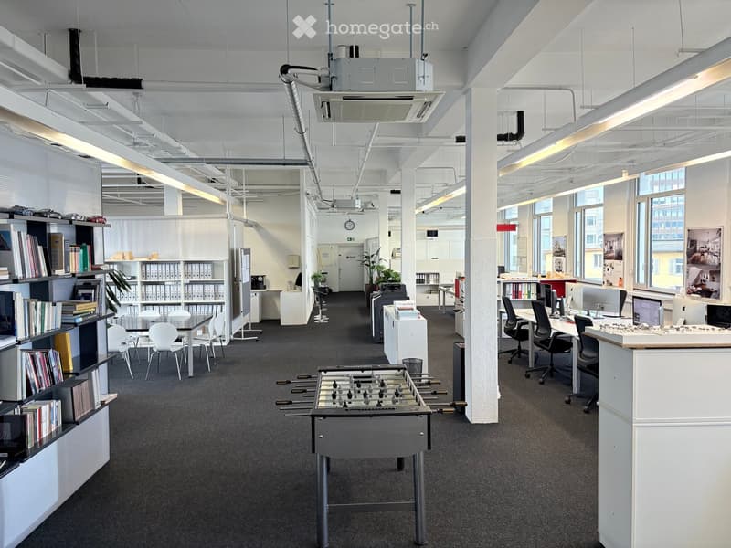 Open office layout with desks, chairs, bookshelves, foosball table, monitors, cabinets, plants, and large windows.