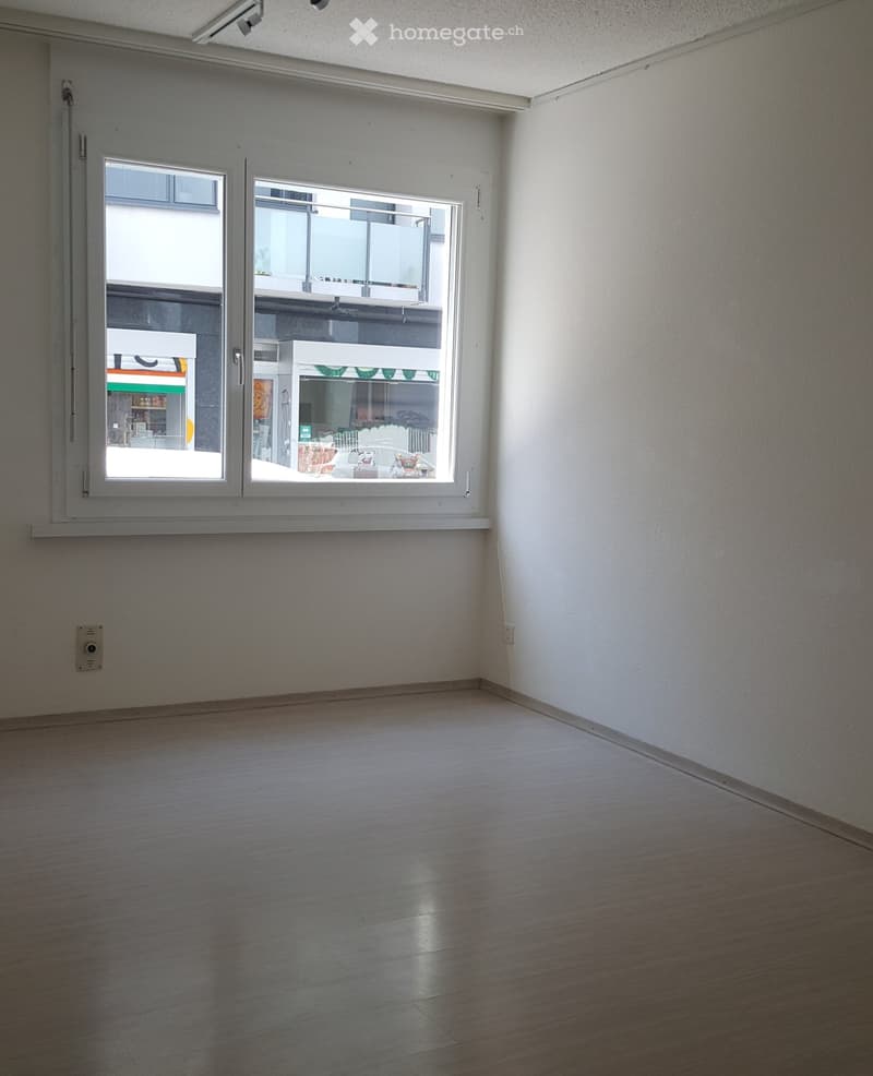 Empty room with white walls, wooden floor, large window, power outlet, and a view of a building outside.