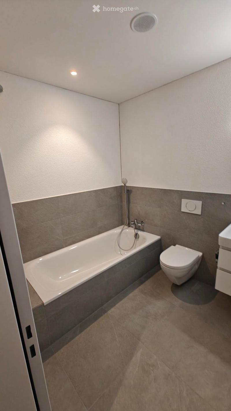 Modern bathroom with a bathtub, toilet, and a faucet, featuring grey tiles and a beige floor.