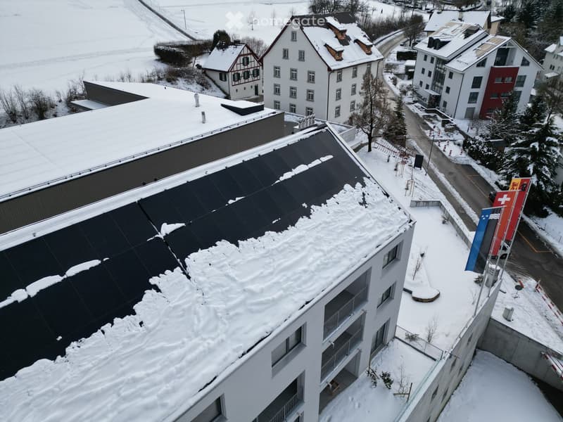 White building with balconies, solar panels on the roof covered in snow, surrounded by snow-covered landscape