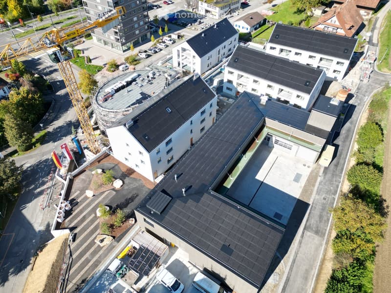 Aerial view of a building construction site, with several buildings under construction, solar panels on the roofs, a crane, and cars parked in the area.