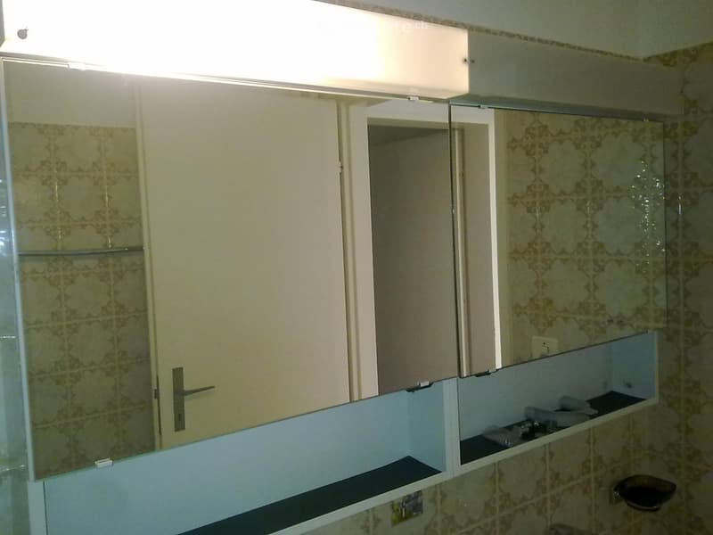 Bathroom mirror with storage shelves, tiled wall behind it, an open white door and a doorframe with another door behind it