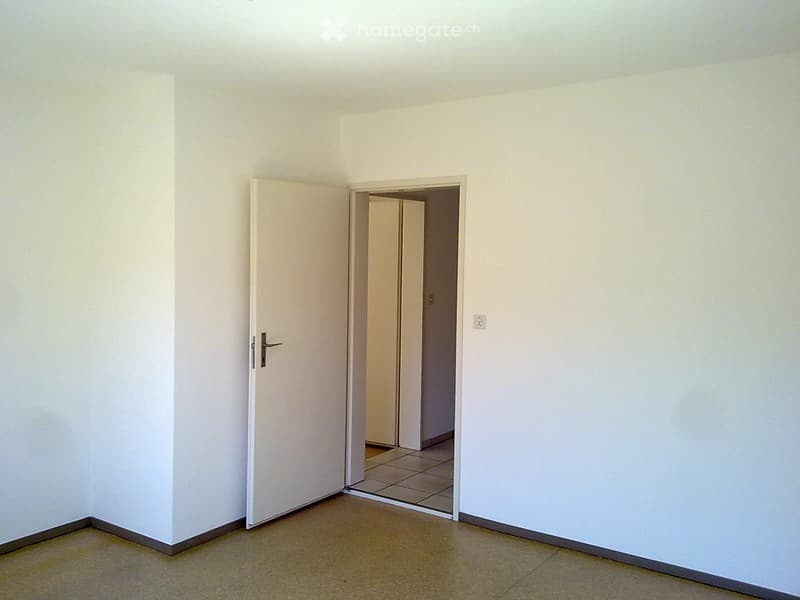 Empty room with white walls and tiled floor, an open door leading to another room, power outlet on the wall.