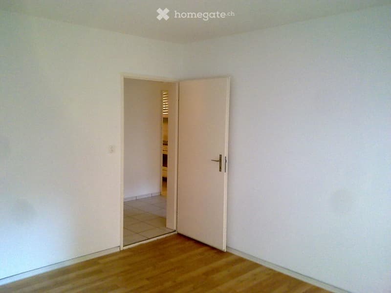 Empty room with wooden floor and white walls. The room has an open door leading to a tiled floor.