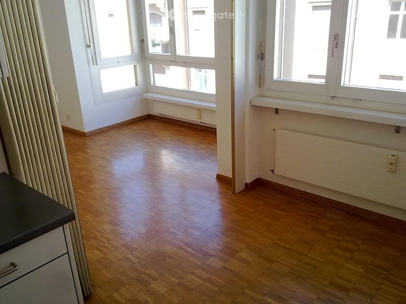 Empty room, wooden floor, white walls, windows with bench, radiator, curtains