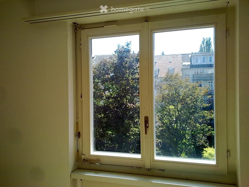 Room with large window overlooking trees and buildings