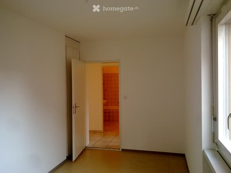 An empty room with white walls and tiled floor, an open door leading to the bathroom.