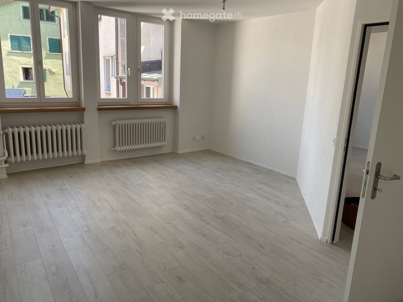 A room with light wooden flooring, two large windows, white walls, and underfloor heating radiators. An open door leading to another room.