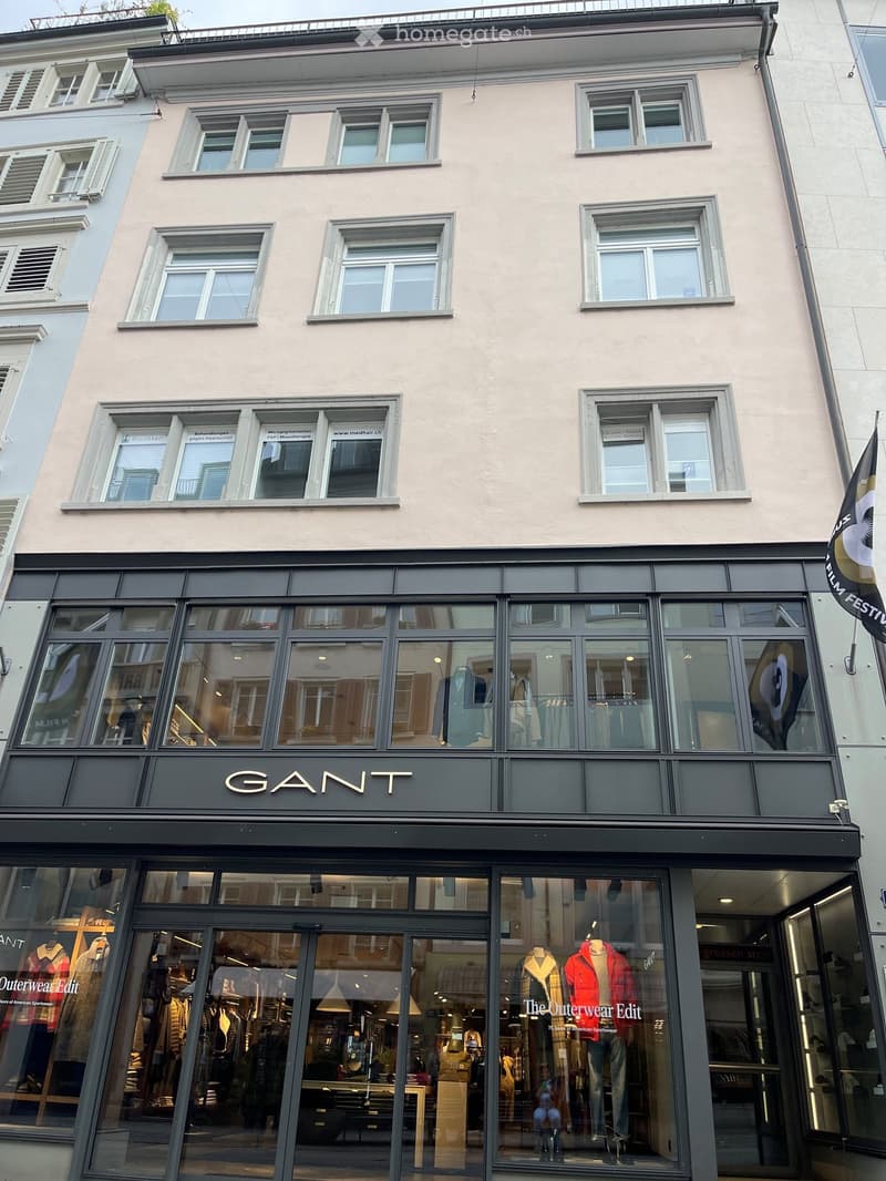 Four-story building with light beige facade, multiple windows, and a storefront labeled "GANT" with large glass windows displaying clothing mannequins and interior store elements.
