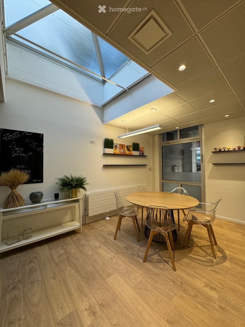 The room features a round wooden dining table with four plastic chairs. There is a glass door with a blind on the side. On the opposite wall, there is a chalkboard and a white shelf with a plant and decorative items. Above the table, there is a hanging la