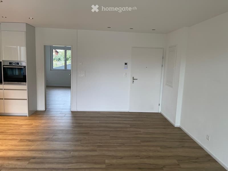 Empty room with wooden flooring, white walls, and built-in cabinets. A doorway leads to another room with a window and tiled floor.