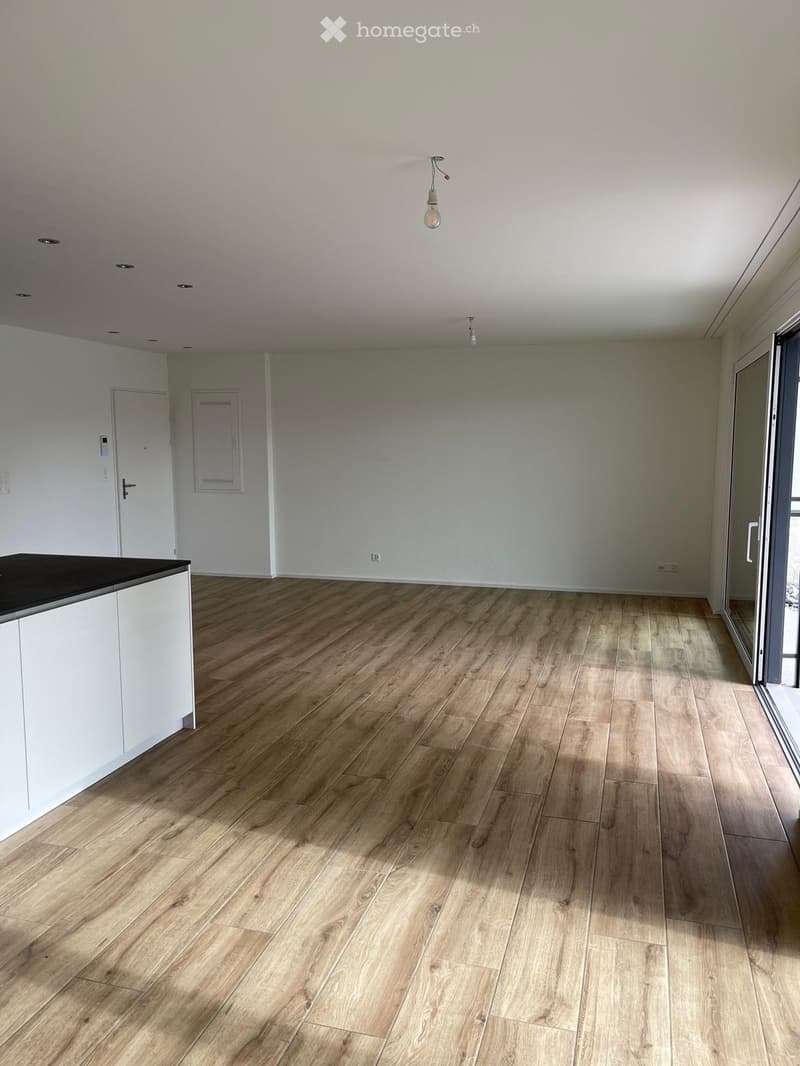 Empty room with light-colored wooden flooring, white walls, and a small white cabinet with a black countertop. There is a door and a window with blinds.
