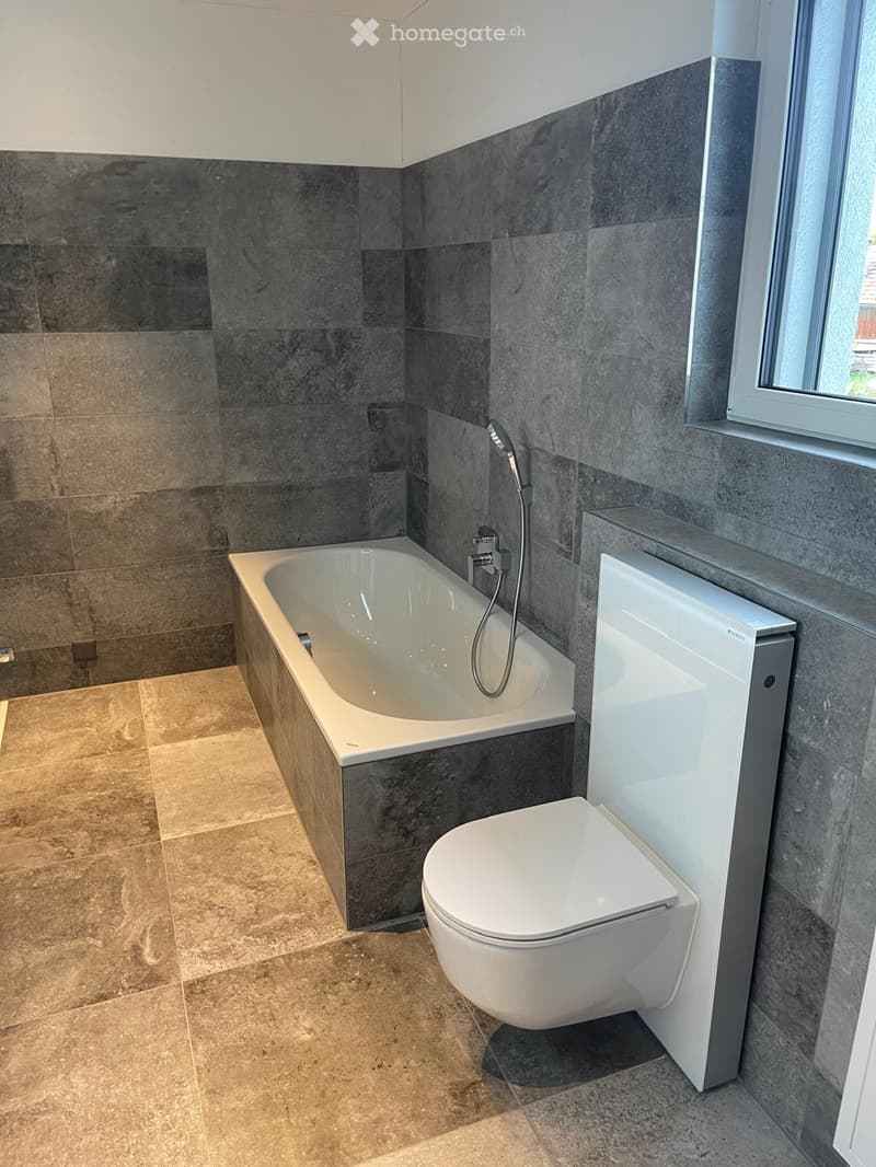 Modern bathroom with a bathtub, toilet, and a handheld showerhead. The walls are tiled in dark grey and light brown, and the floor is tiled in light brown.