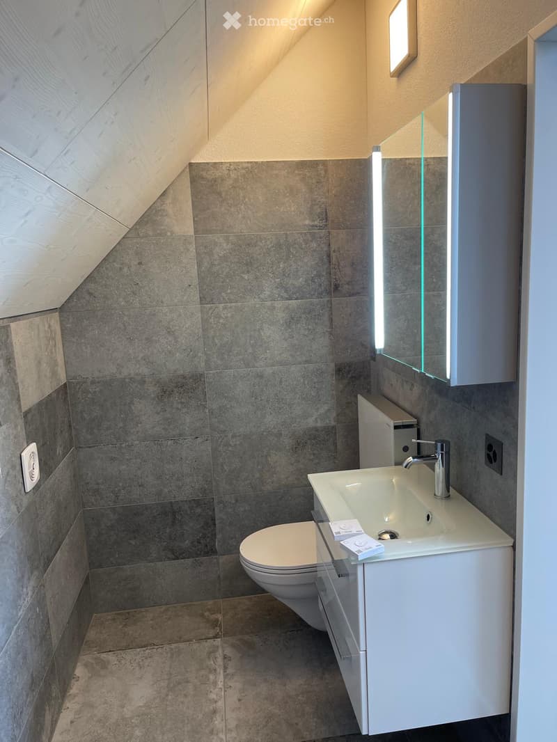 Bathroom. Modern bathroom with grey tiled walls, white sink, chrome faucet, toilet, and illuminated mirror.