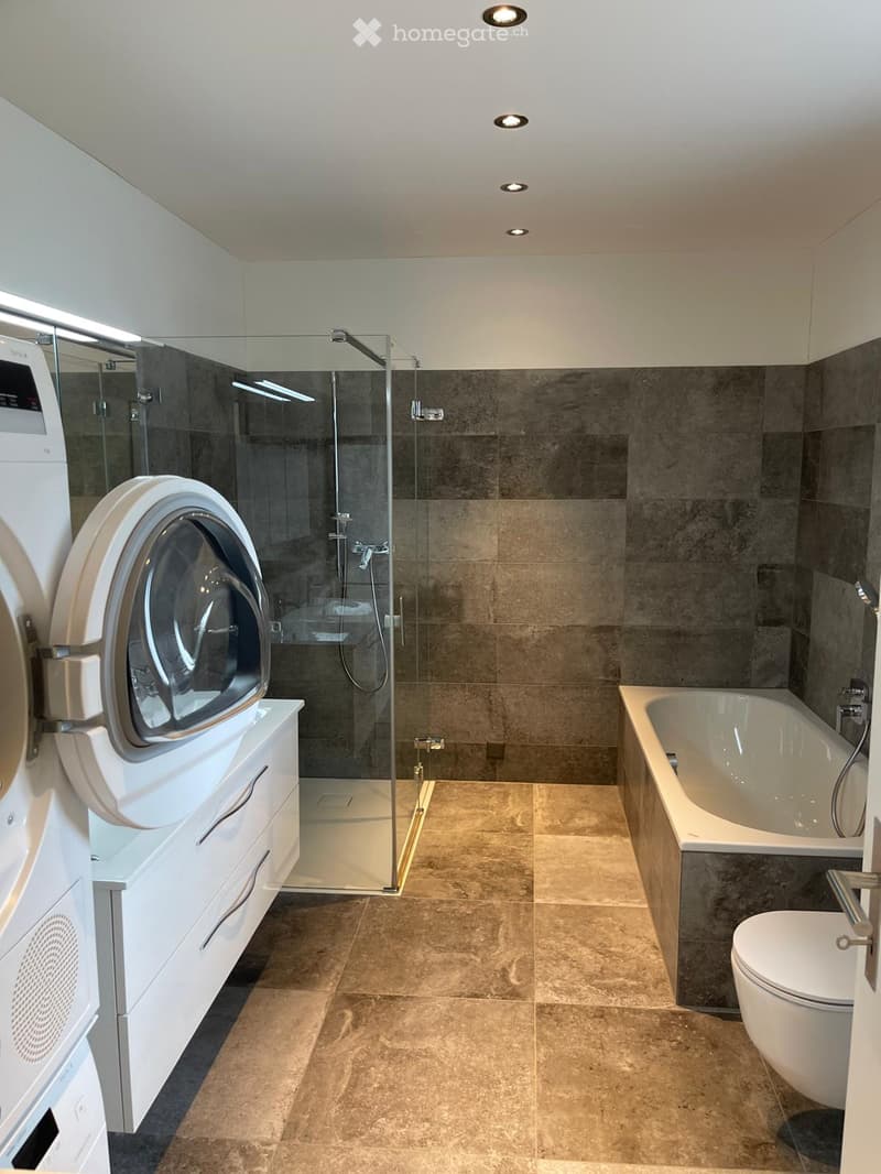 Modern bathroom with a bathtub, toilet, glass-enclosed shower, and a washing machine on a white cabinet.