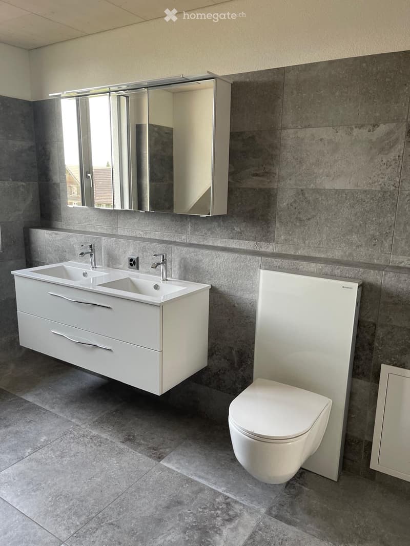 Bathroom. Modern bathroom with a double sink vanity, sleek white cabinetry, a large mirror, and a toilet. The walls are tiled in a grey stone pattern, and the floor is covered with matching grey tiles.
