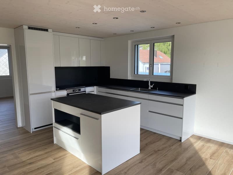Kitchen. Modern kitchen with white cabinets, black countertops, and a central island with an integrated oven. The space features wooden flooring and large windows offering a view of the exterior.