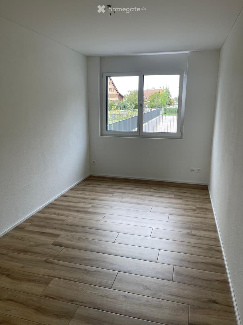 Empty room with light wooden flooring and white walls, featuring a large window with a view of the outside area.