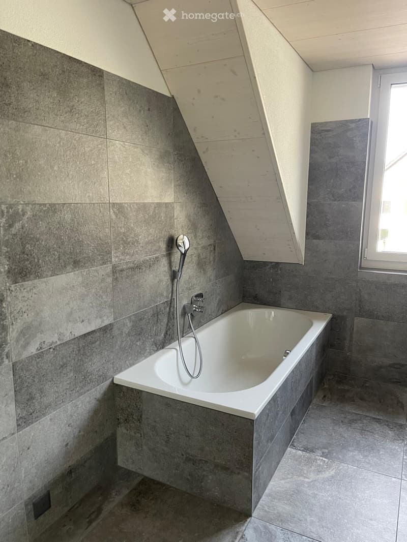 Bathroom. Modern bathroom with a white bathtub, grey tiled walls, and a showerhead attached to the wall.