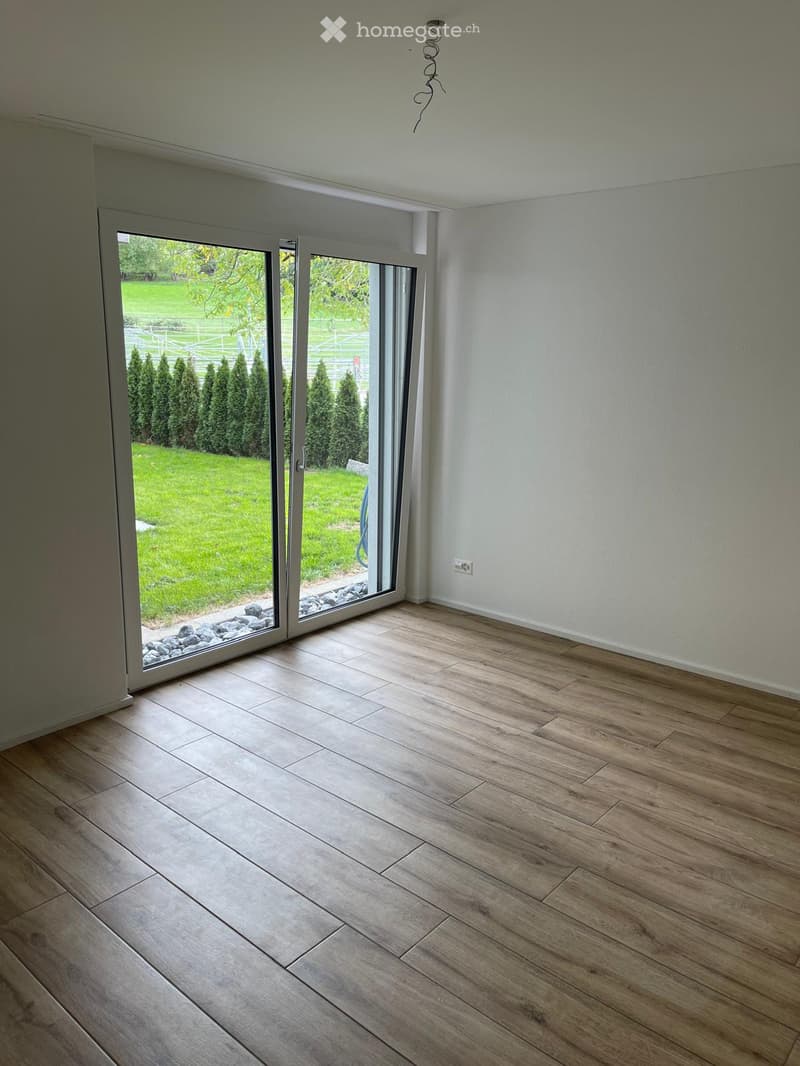 Empty room with wooden flooring, large sliding glass doors leading to a green lawn and trees, white walls, and visible electrical outlet.