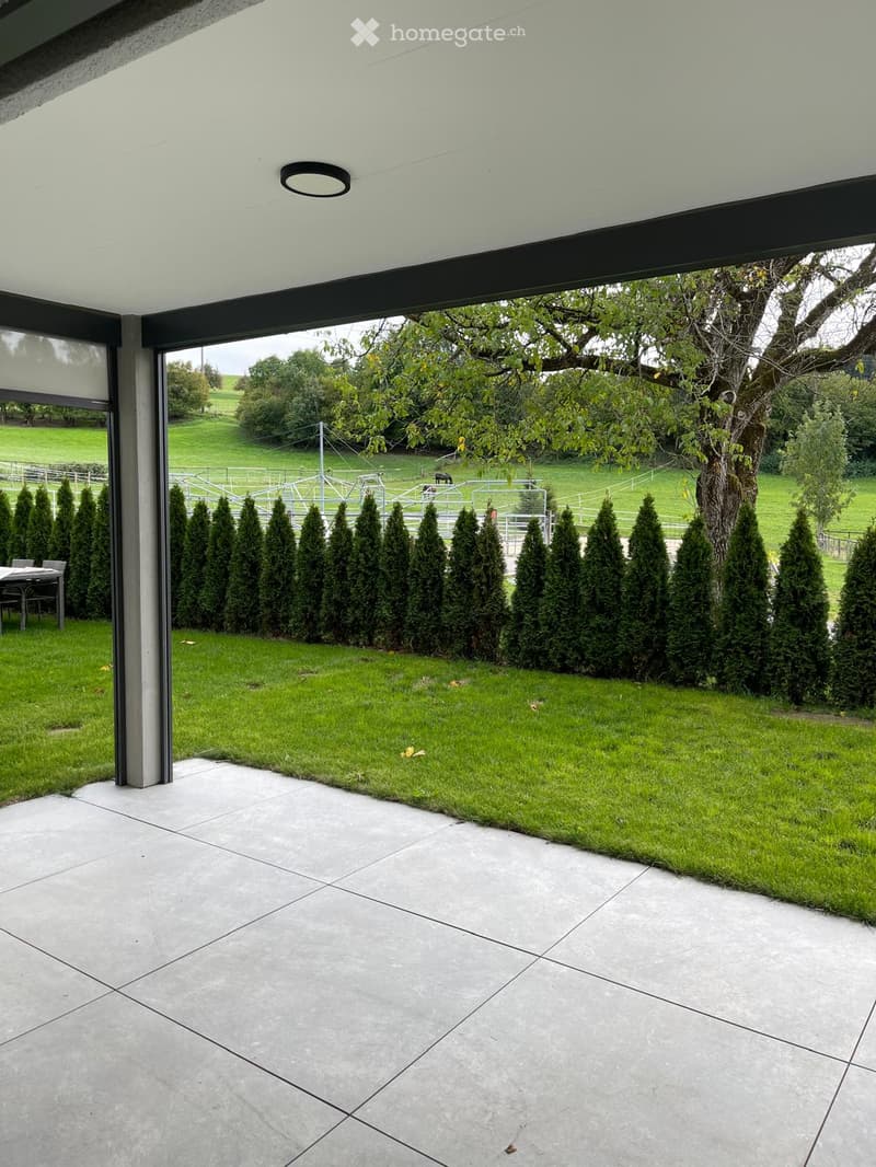 Modern balcony with large glass walls, tiled floor, and a view of a well-maintained lawn and trees.