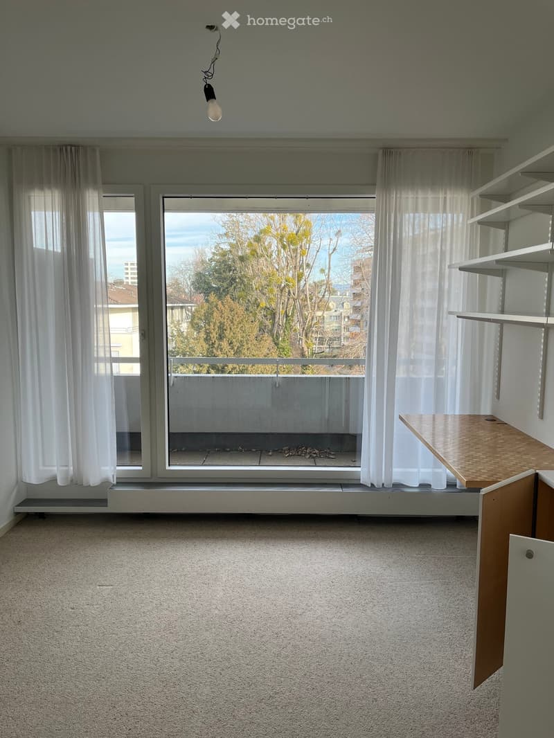 empty room, large window with access to balcony, white curtains, wooden floor, desk on the right