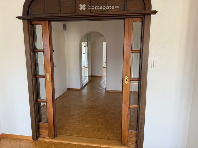 Open hallway with arched entrance, wooden flooring, white walls, visible a door to the left