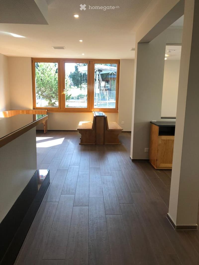 An empty room with wooden benches, a desk, and a large window offering a view of the outdoors.