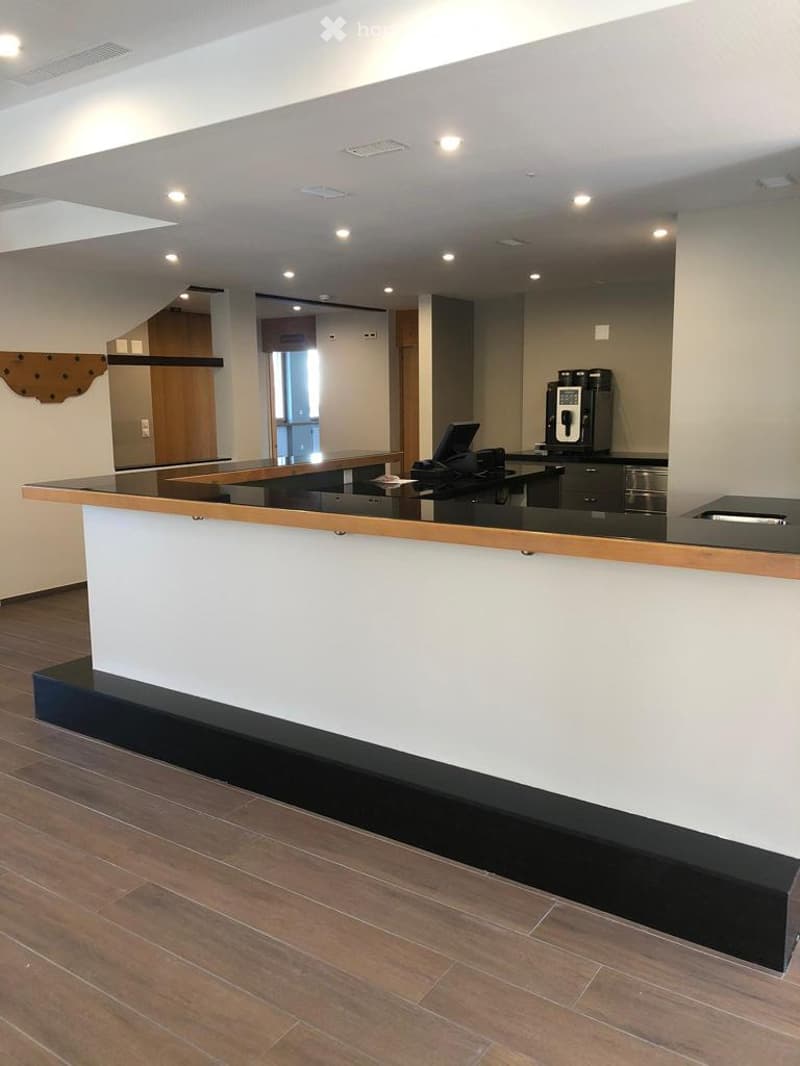 reception area with a black countertop, wooden edges, a black computer and a coffee machine, white walls, a wooden entrance door with a mounted sign, a large window, wooden flooring